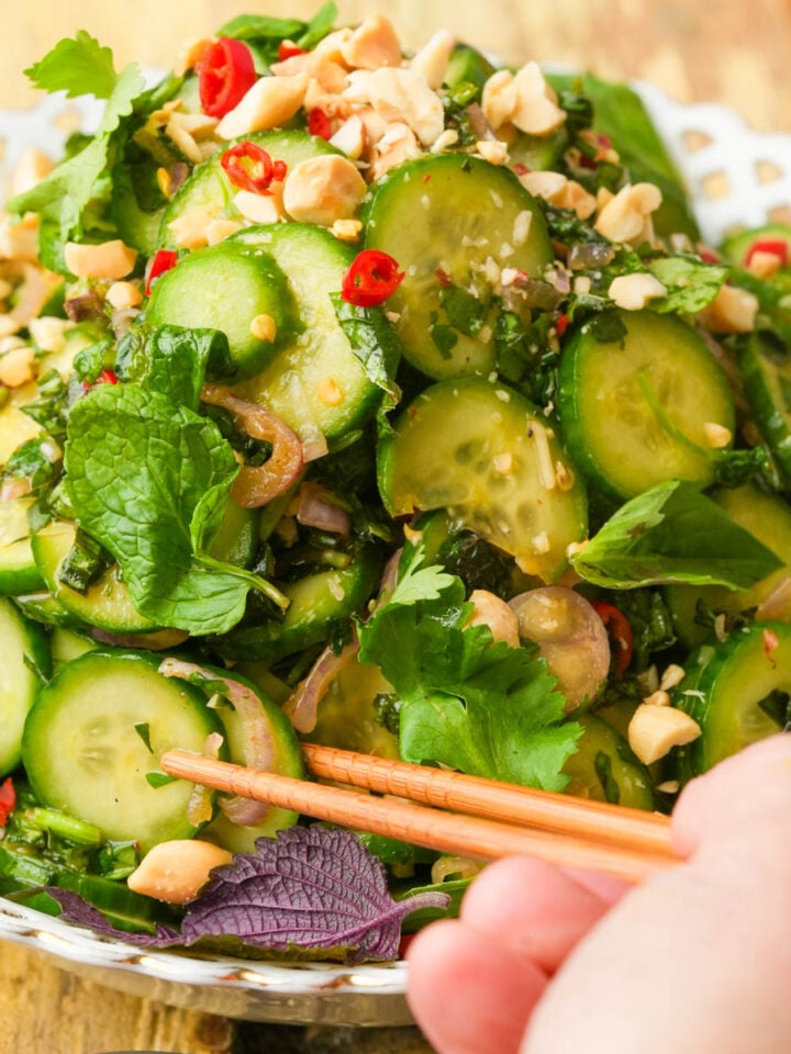 A hand holding chopsticks reaches for a fresh cucumber salad garnished with chopped peanuts, herbs, red chili slices, and leafy greens, served in a white decorative bowl.