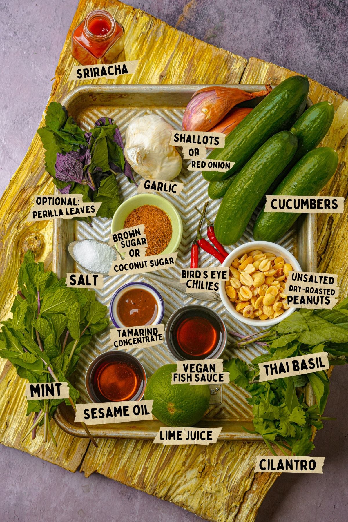 A tray with ingredients for Thai cucumber salad: sriracha, shallots or red onion, garlic, cucumbers, brown or coconut sugar, bird's eye chilies, unsalted peanuts, salt, tamarind, vegan fish sauce, lime juice, Thai basil, cilantro, mint, sesame oil, and perilla leaves.