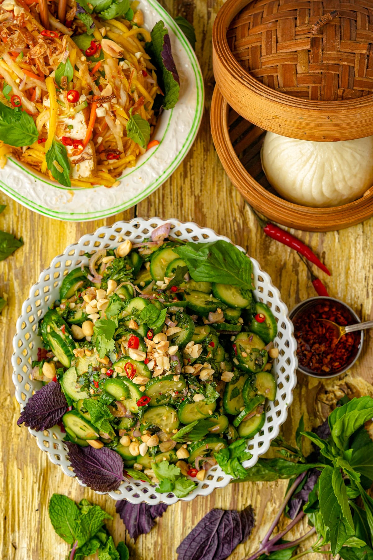 A vibrant spread of Asian dishes on a wooden table, featuring a cucumber salad topped with peanuts and herbs, a yellow mango salad, a bamboo steamer with a white bun, and fresh herbs scattered around.