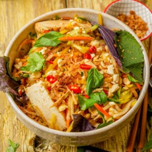 A bowl of Vietnamese noodle salad with tofu, fresh herbs, peanuts, chili slices, shredded vegetables, and crispy onions on a wooden table. Chopsticks and a small bowl of toppings are nearby.