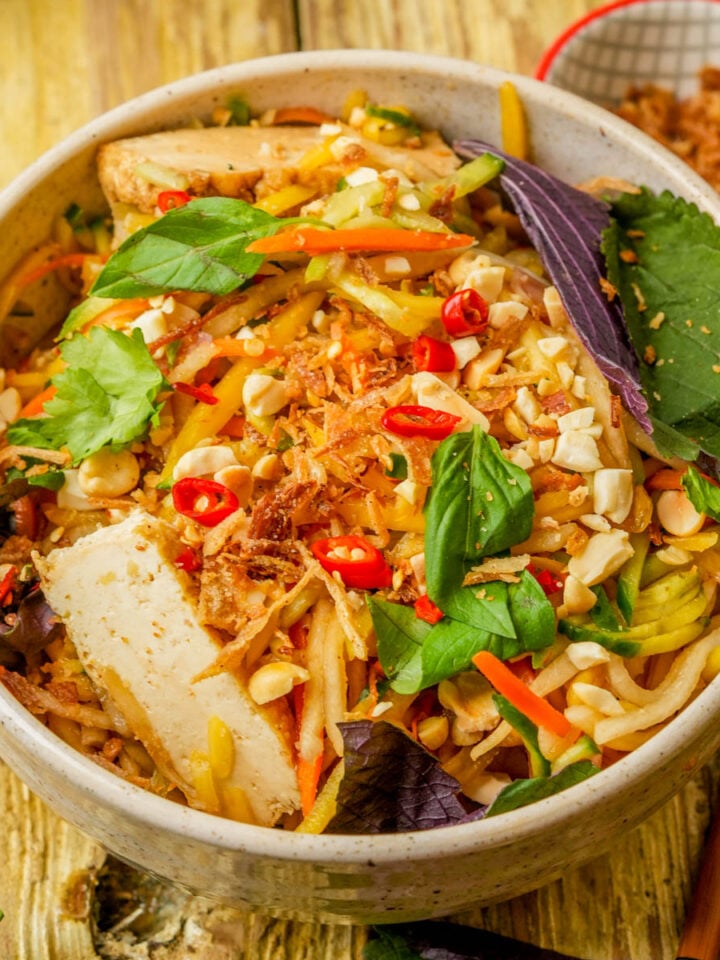 A bowl of Vietnamese noodle salad with tofu, fresh herbs, peanuts, chili slices, shredded vegetables, and crispy onions on a wooden table. Chopsticks and a small bowl of toppings are nearby.