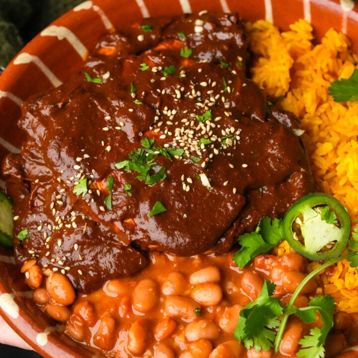 A plate with yellow rice, pinto beans in sauce, and vegan chicken covered in dark mole poblano sauce, garnished with sesame seeds, fresh cilantro, and slices of jalapeño.
