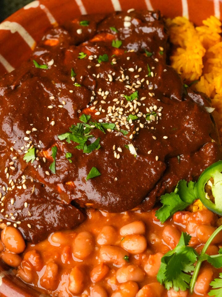 A plate with yellow rice, pinto beans in sauce, and vegan chicken covered in dark mole poblano sauce, garnished with sesame seeds, fresh cilantro, and slices of jalapeño.