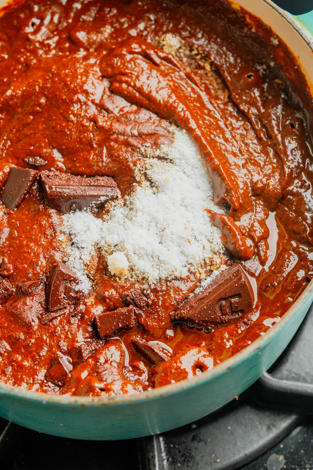 A close-up of a pot on a stove containing a thick, red mole poblano sauce with chunks of dark chocolate and a mound of white salt or sugar being added.
