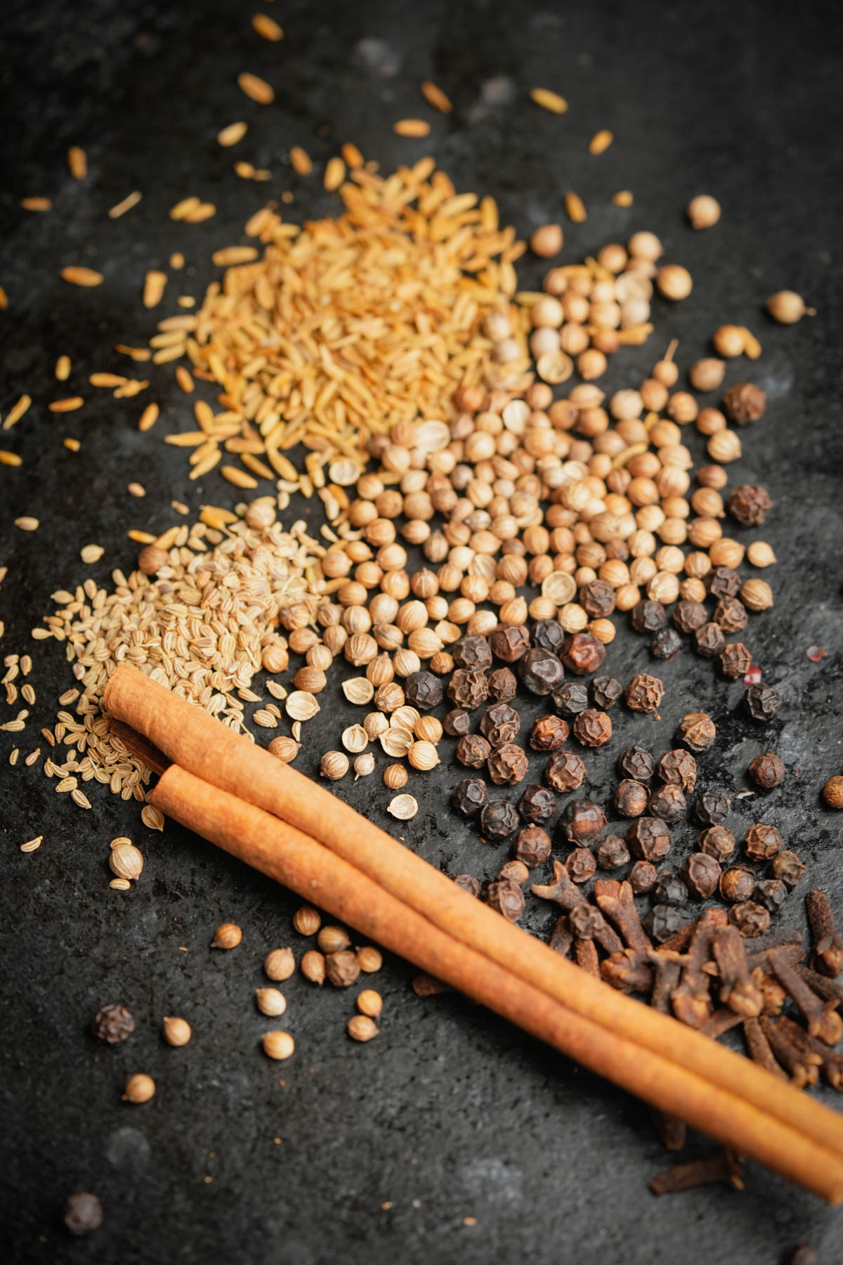 A flat lay of whole spices on a dark surface, including coriander seeds, black peppercorns, cloves, rice grains, and two cinnamon sticks.