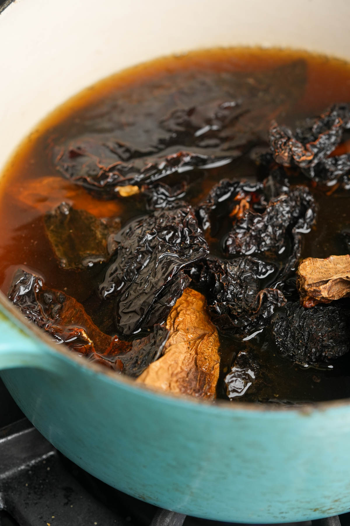 A close-up of dried chilies ginger simmering in dark broth inside a light blue pot on a stove.