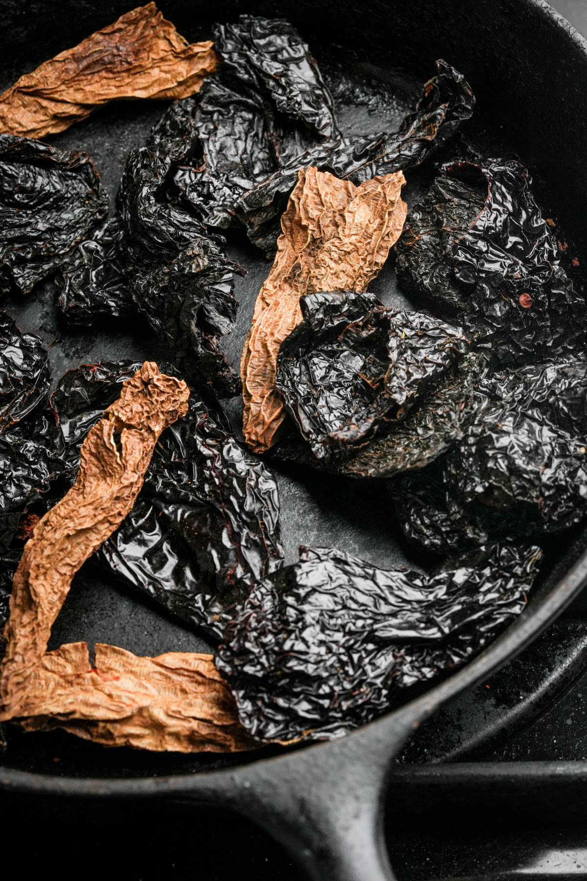 Dried chipotle and ancho peppers in a black cast iron skillet. The wrinkled, dark peppers contrast with the lighter, reddish-brown strips on the pan's surface.