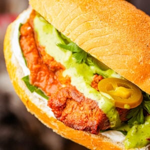 Close-up of a vegan carnitas torta with green sauce, sliced jalapeños, fresh cilantro, and mayonnaise on a toasted bun.