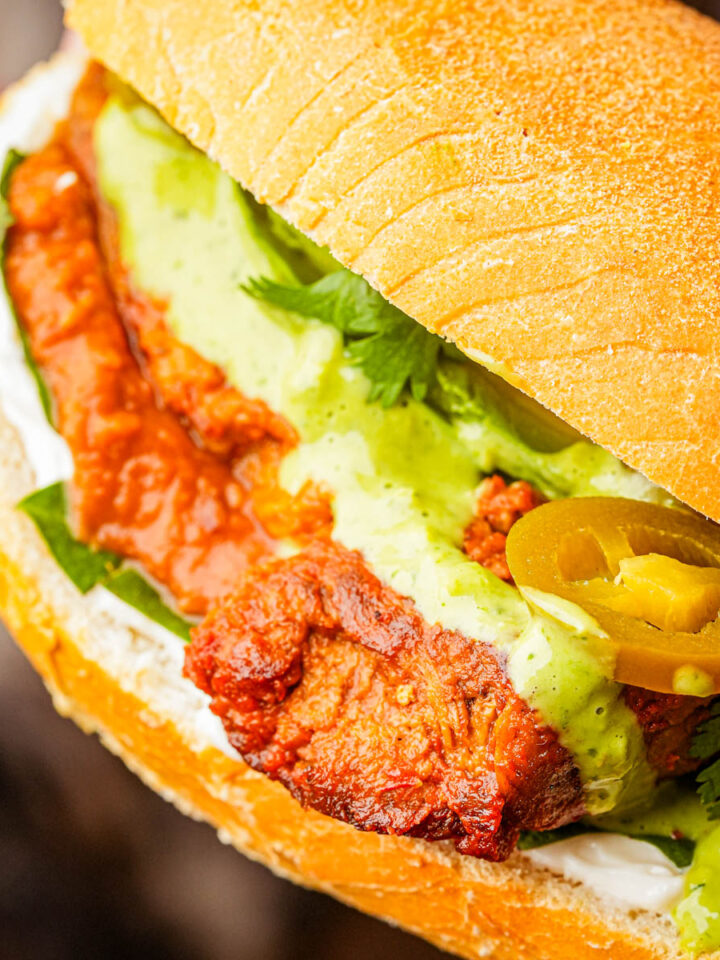 Close-up of a vegan carnitas torta with green sauce, sliced jalapeños, fresh cilantro, and mayonnaise on a toasted bun.