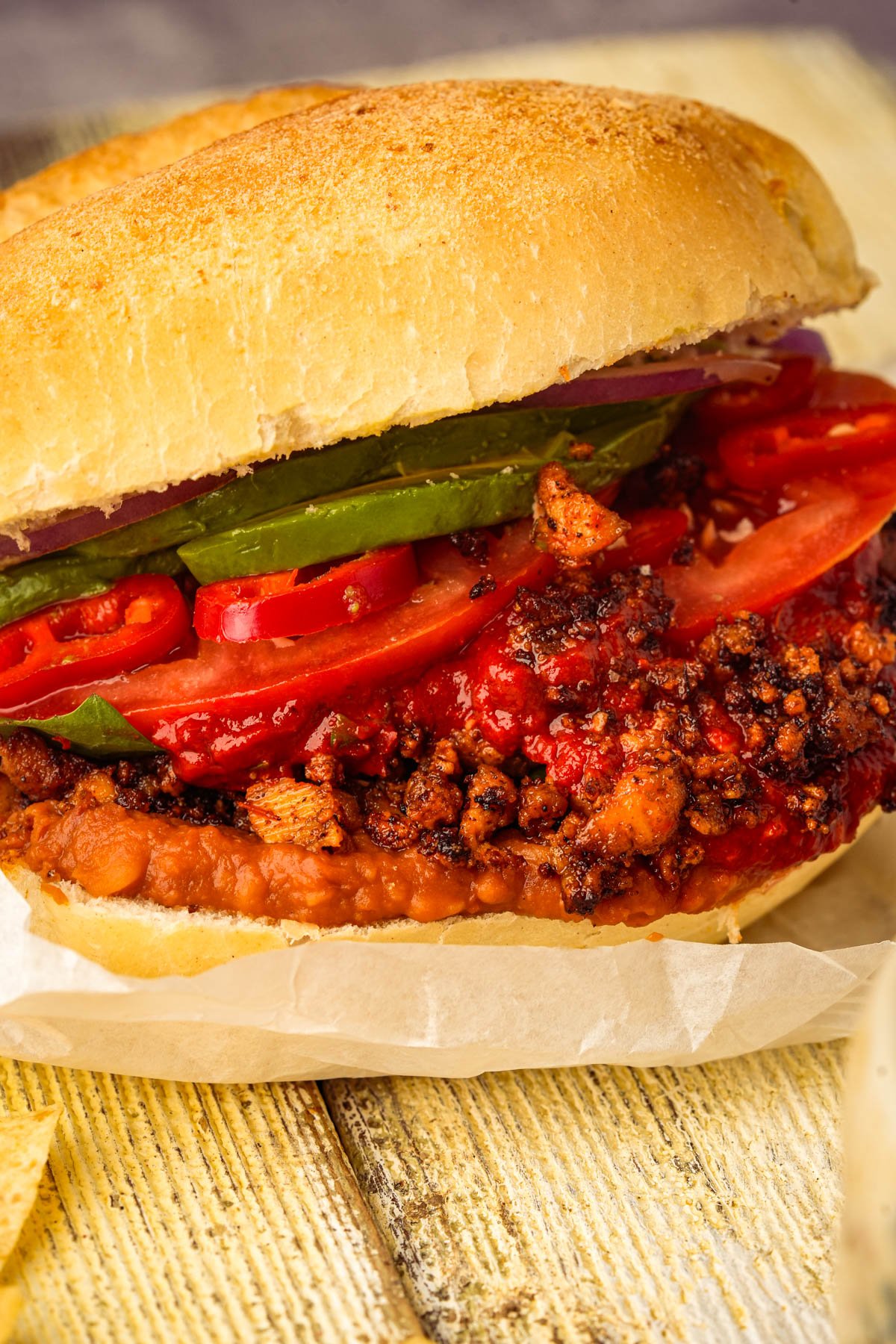A Mexican vegan chorizo torta with crusty bread, refried beans, seasoned ground tofu meat, tomato slices, jalapeño peppers, and red chili slices, served on parchment paper on a rustic wooden surface.