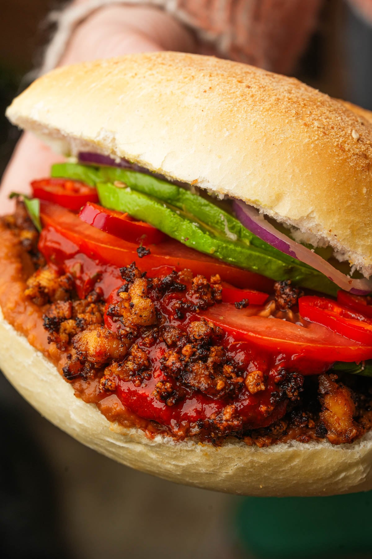 A hand holding a vegan torta de chorizo filled with s sliced tomatoes, avocado, red onion, and sauce on a round bread roll.