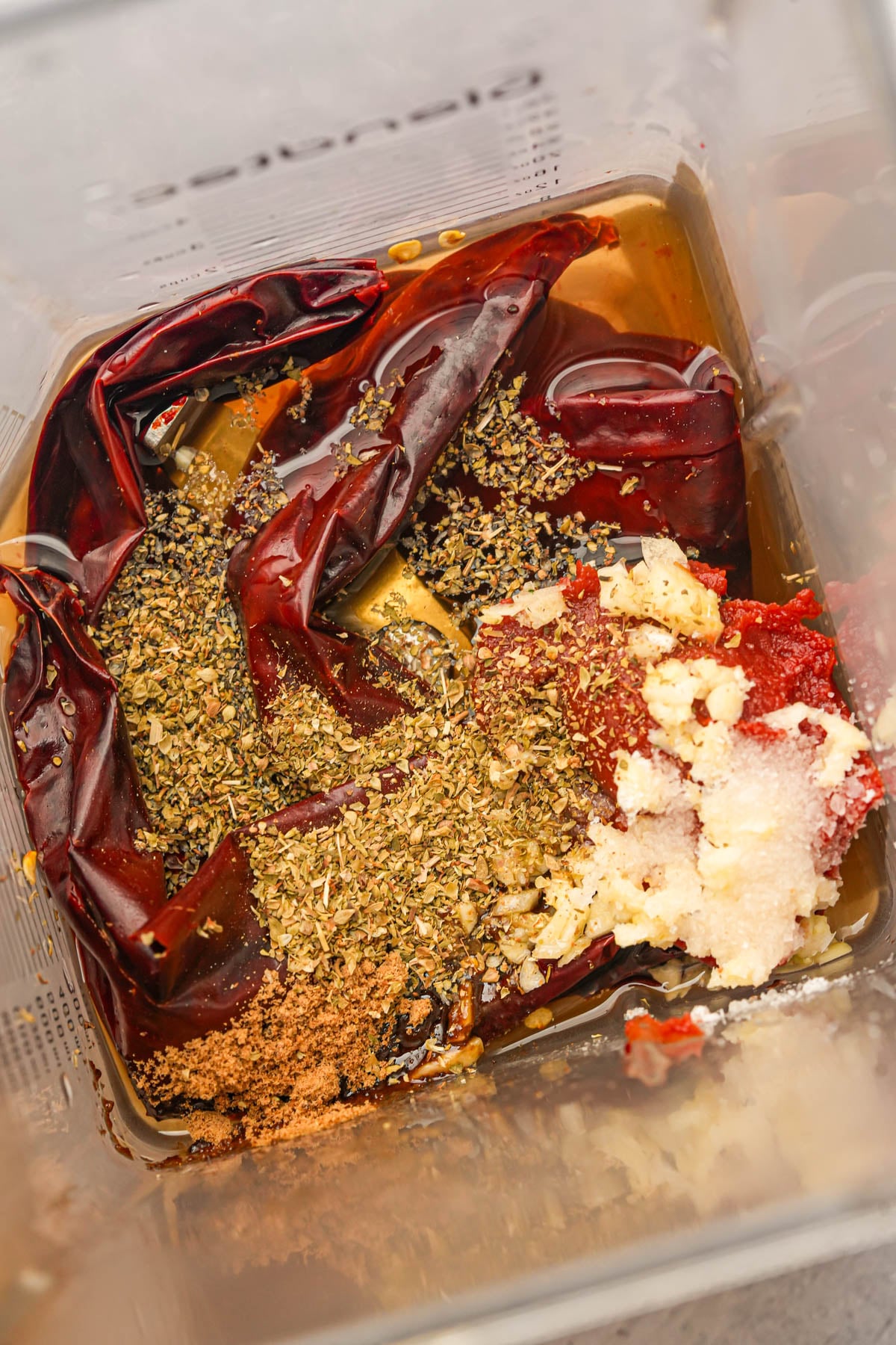 A blender container filled with dried red chilies, oregano, cumin, garlic, sugar, and a dark liquid, ready to be blended into a sauce.