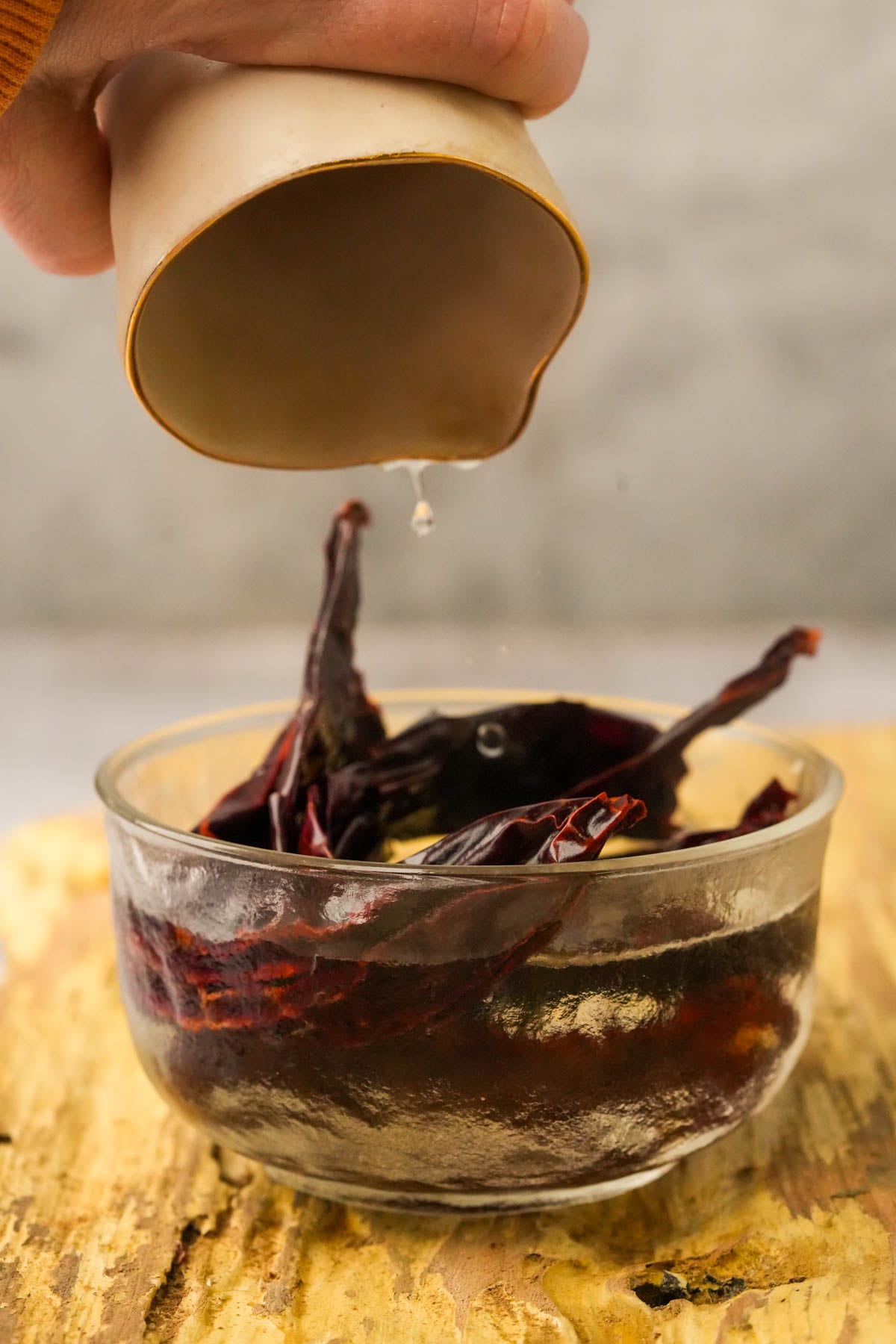 A hand pours liquid from a small cup into a glass bowl filled with dried red chilies soaking in water, placed on a wooden surface.