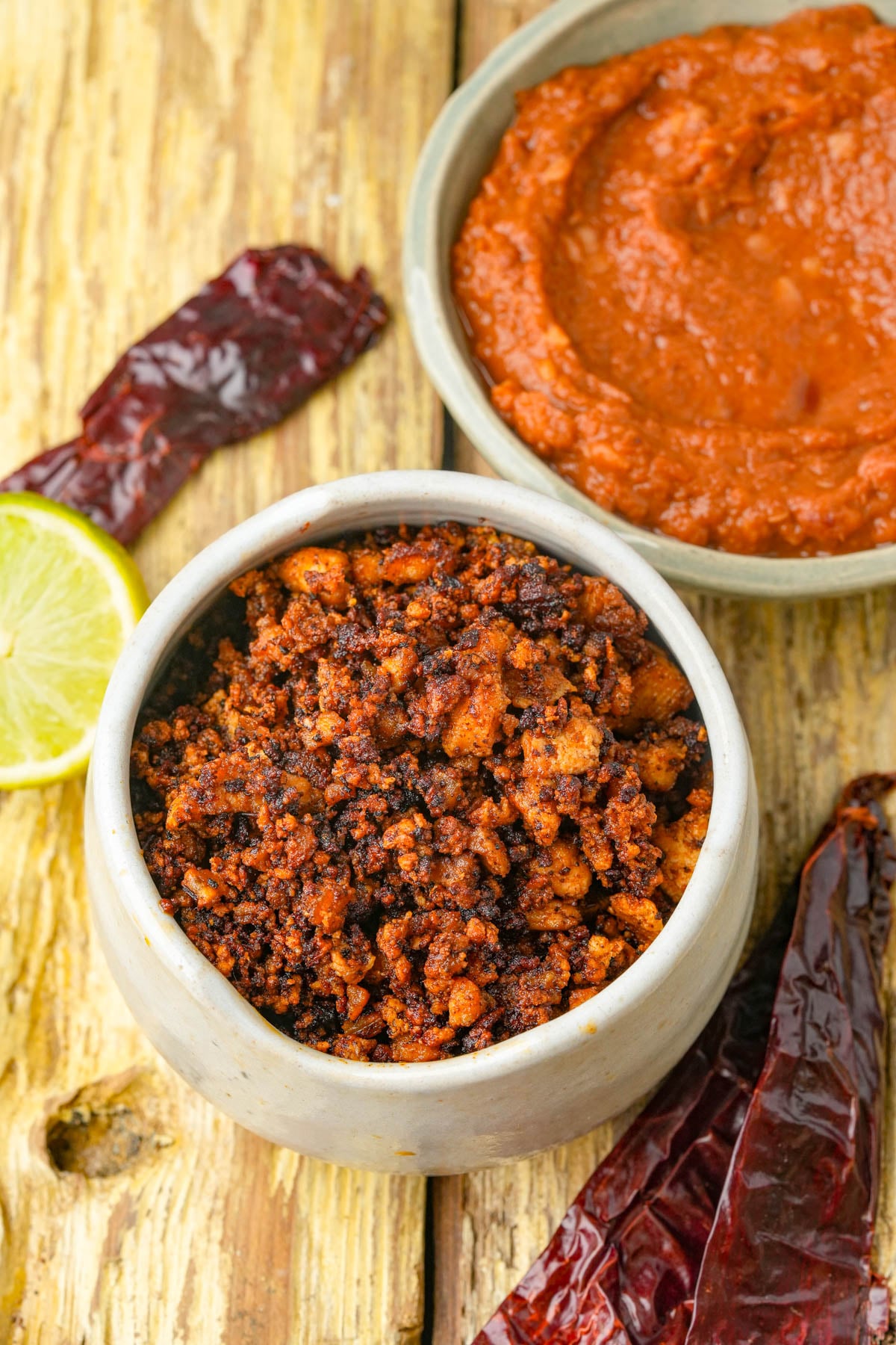 A small ceramic bowl filled with spicy vegan chorizo sits on a rustic wooden surface, surrounded by dried red chilies, a lime slice, and a bowl of refried beans.