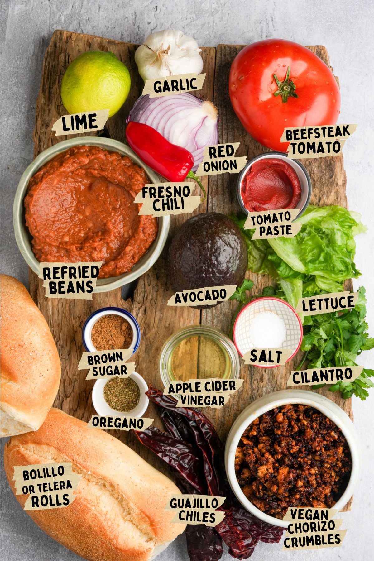 A wooden board displays ingredients for vegan torta recipe: lime, garlic, beefsteak tomato, red onion, tomato paste, lettuce, cilantro, salt, avocado, fresno chili, refried beans, brown sugar, oregano, apple cider vinegar, guajillo chiles, vegan chorizo crumbles, and bolillo rolls.