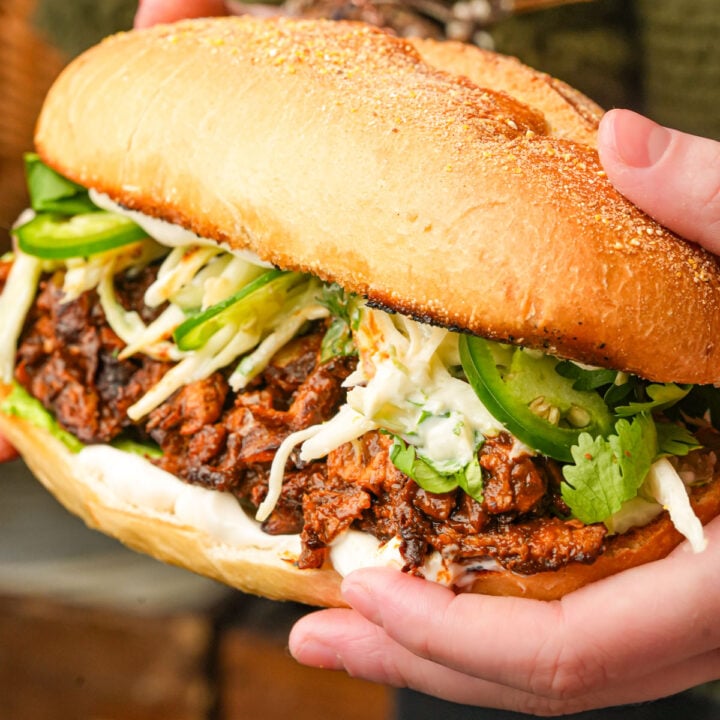 A person holds a large vegan chicken mole torta filled with soy curls, sliced jalapeños, cilantro, slaw, and sauce on a toasted bolillo.