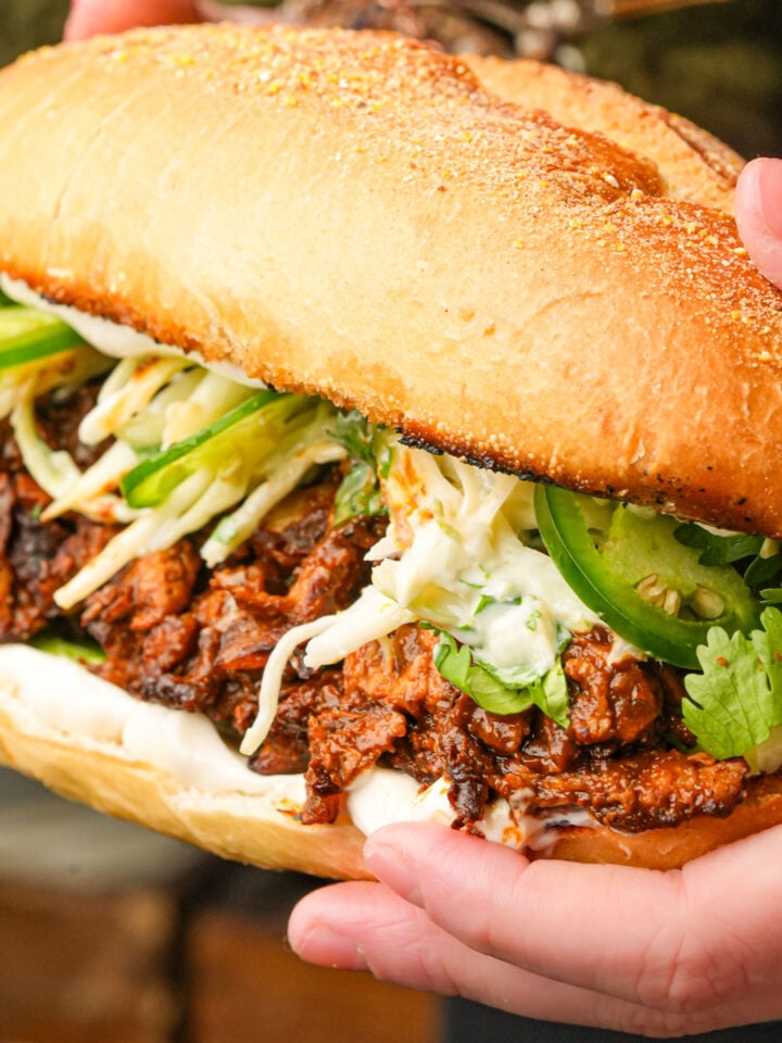 A person holds a large vegan chicken mole torta filled with soy curls, sliced jalapeños, cilantro, slaw, and sauce on a toasted bolillo.