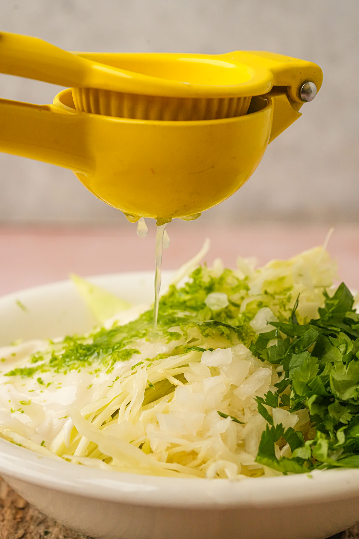 A yellow citrus squeezer is squeezing lime juice onto a bowl filled with shredded cabbage, vegan sour cream, onions, and chopped cilantro.
