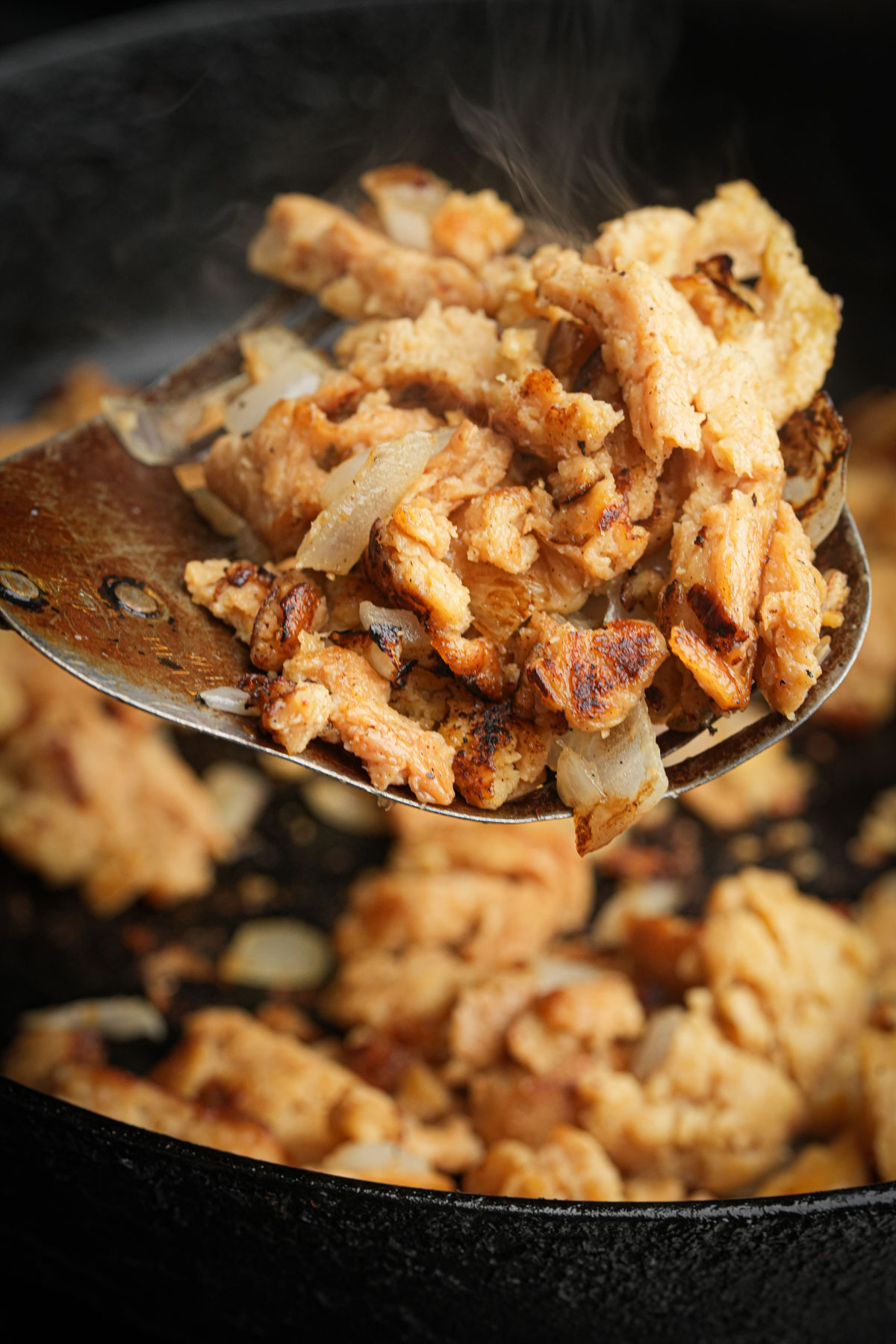 A metal spatula lifts a portion of browned, cooked soy curls with onions from a black skillet, showing steam rising from the hot food.