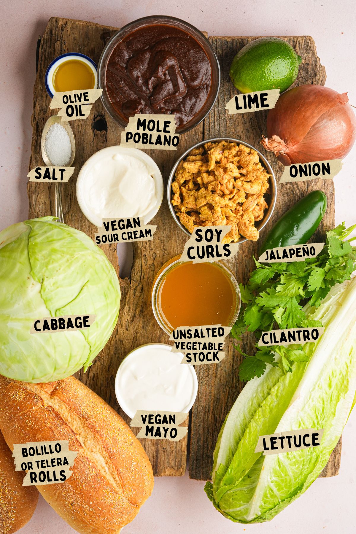 Ingredients for a vegan chicken mole torta recipe arranged on a wooden board, including cabbage, lettuce, bolillo rolls, soy curls, mole poblano, vegan mayo, vegan sour cream, onion, jalapeño, cilantro, lime, salt, olive oil, and vegetable stock.