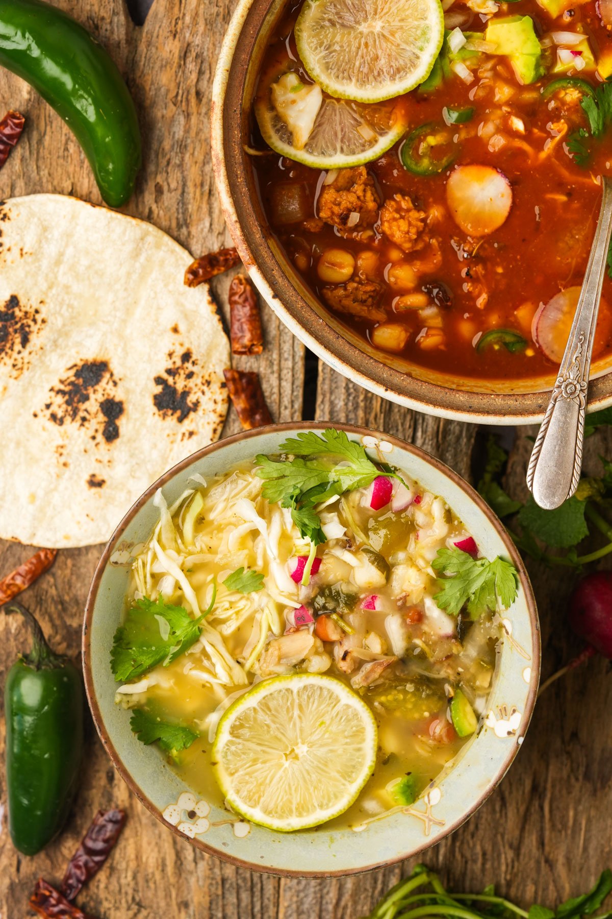 Two bowls of vegan Mexican pozole—one red and one green—topped with lime slices, radish, and cilantro, sit on a rustic wooden table beside a toasted corn tortilla, jalapeños, and dried chilies.