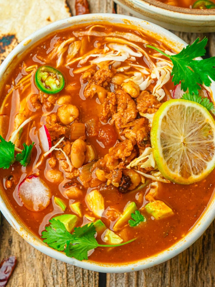 A bowl of vegan pozole rojo with shredded soy curls, beans, avocado chunks, sliced radish, jalapeño, cabbage, cilantro, and a lime slice, served on a rustic wooden table with a tortilla and fresh jalapeño nearby.