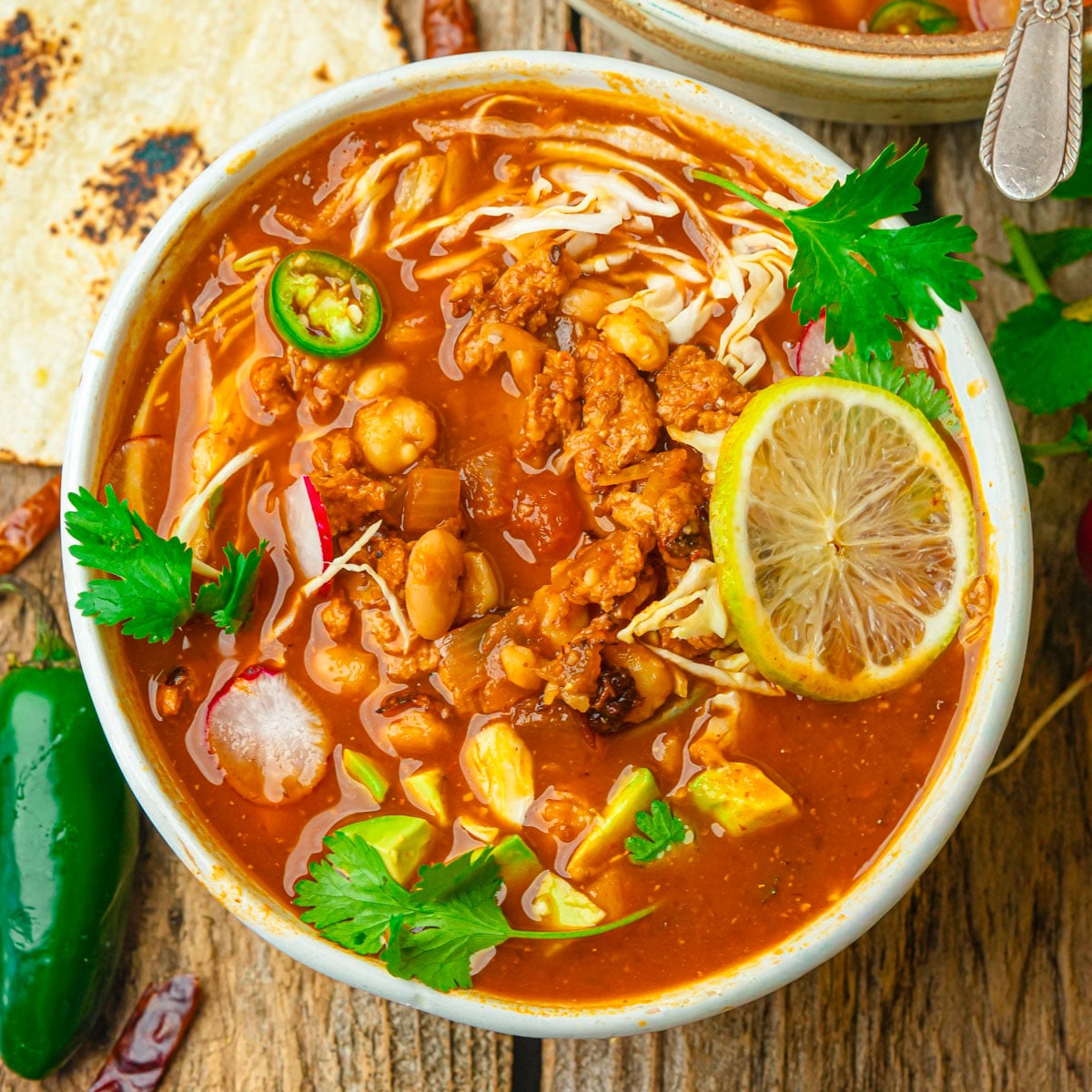 A bowl of vegan pozole rojo with shredded soy curls, beans, avocado chunks, sliced radish, jalapeño, cabbage, cilantro, and a lime slice, served on a rustic wooden table with a tortilla and fresh jalapeño nearby.