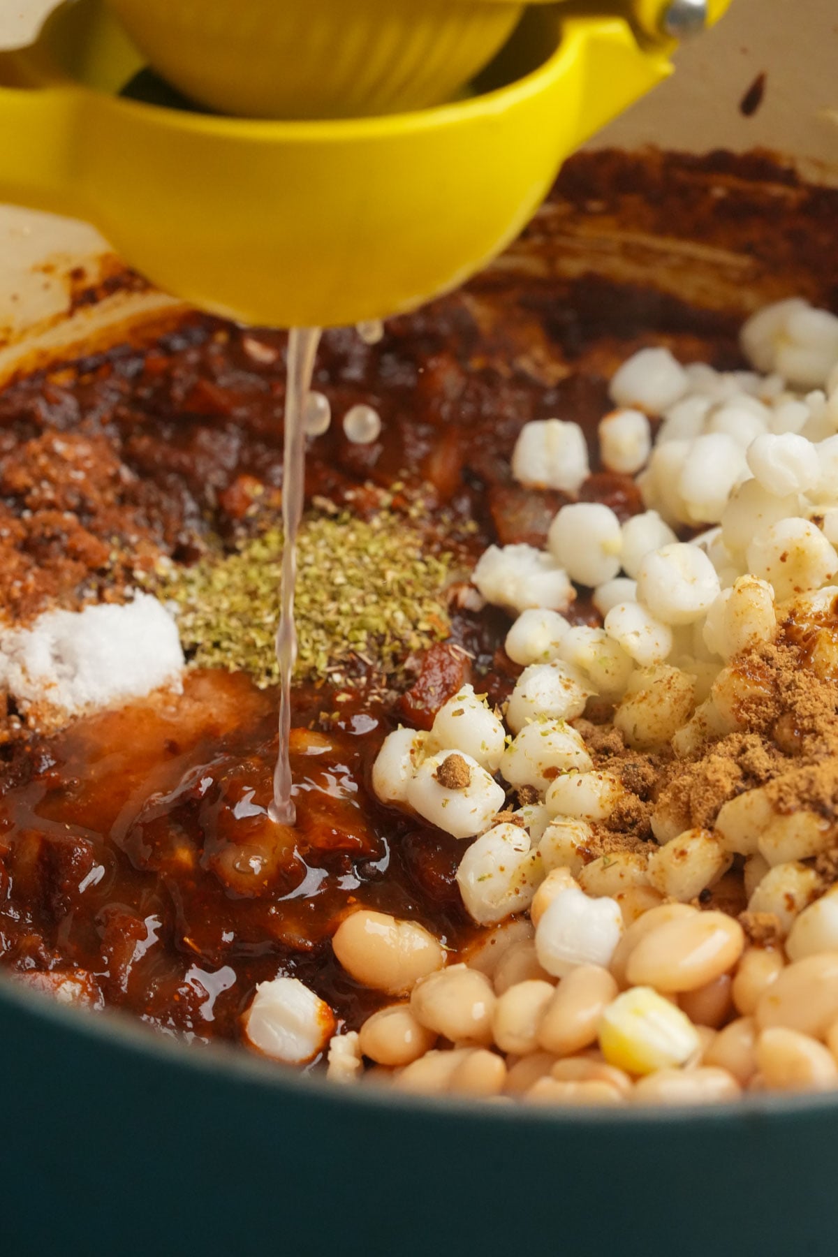 A pot filled with beans, hominy, spices, and chile sauce, while fresh lime juice is being squeezed in with a yellow citrus press.