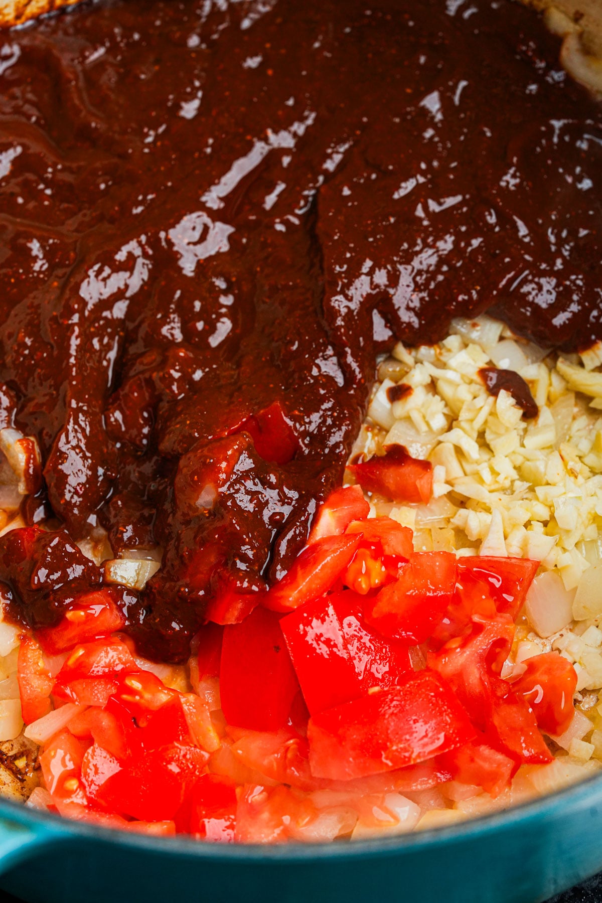 Chopped garlic, onions, and fresh diced tomatoes in a pot, topped with a thick layer of dark red chile puree, ready to be mixed and cooked together.