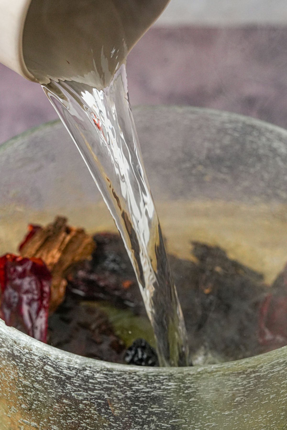 Boiling water is being poured from a white pitcher into a glass bowl containing dried chilies.