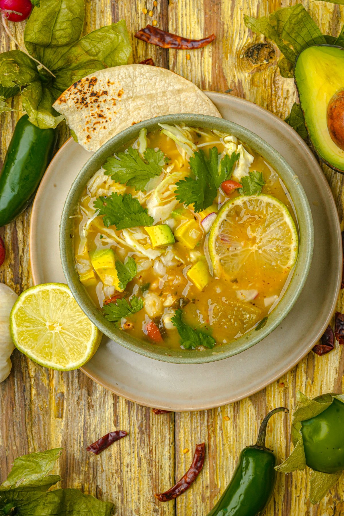A bowl of vegan pozole verde topped with cilantro, avocado chunks, and a lime slice, served with toasted tortillas on a rustic wooden table, surrounded by fresh chili peppers, tomatillos, garlic, and a halved avocado.