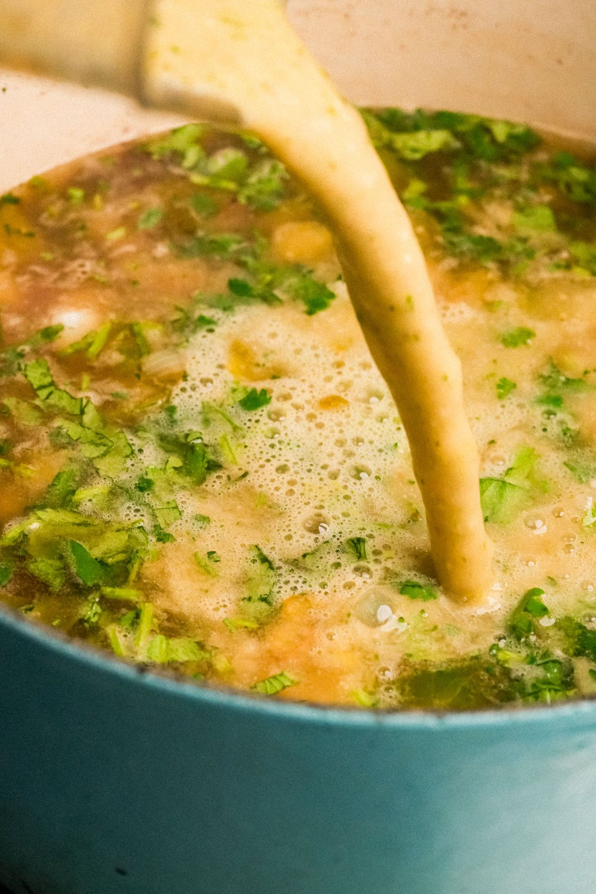 A blender of pureed base is being poured into a pot of vegan pozole verde filled with chopped herbs and vegetables.