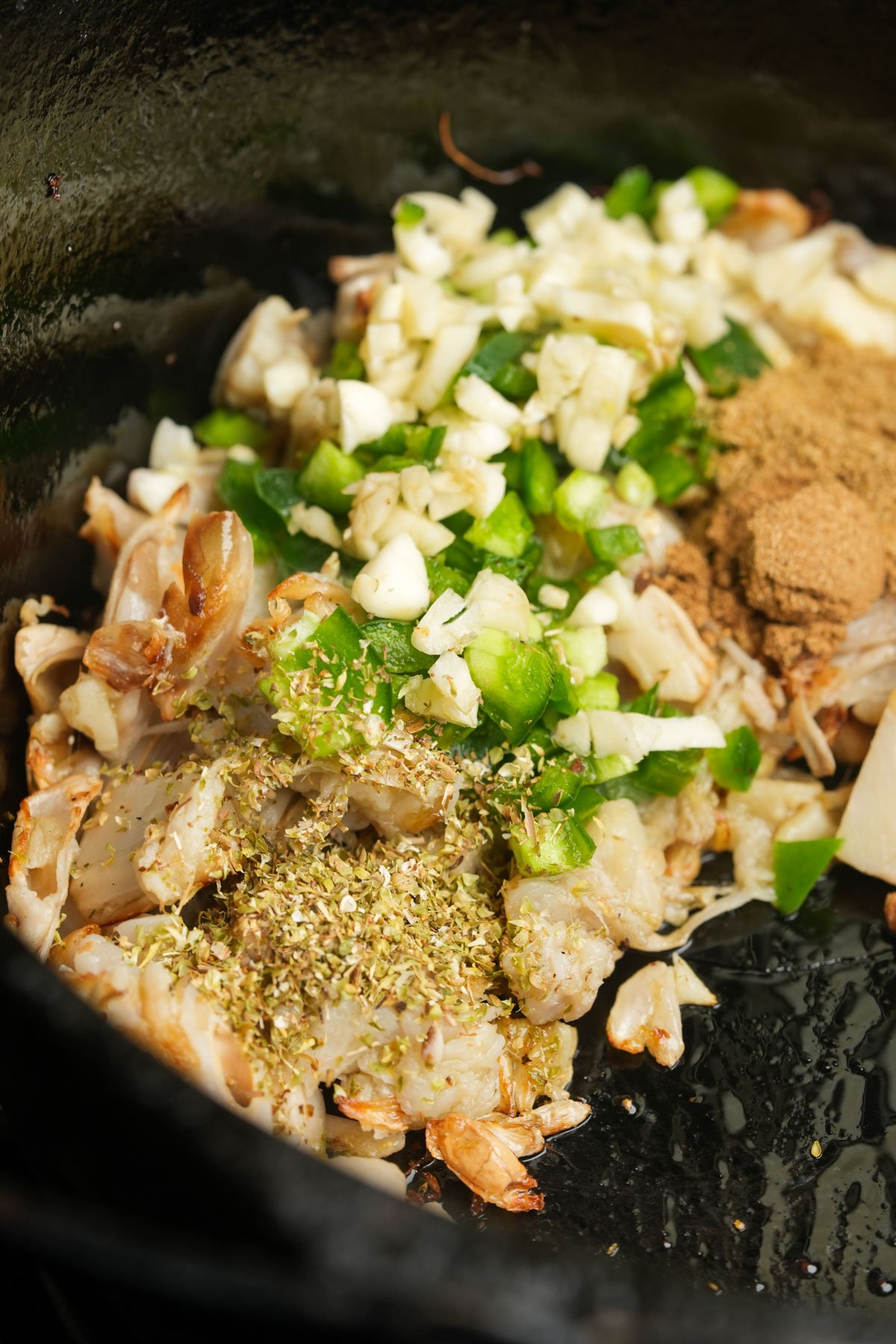 Chopped garlic, green peppers, dried herbs, spices, and cooked jackfruit in a black cast iron skillet, ready to be mixed and cooked.