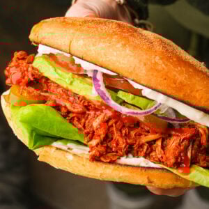 A close-up of a hand holding a large vegan torta adobada filled with shredded barbecue jackfruit, lettuce, avocado slices, tomato, red onion, pickles, and mayonnaise in a toasted bun.