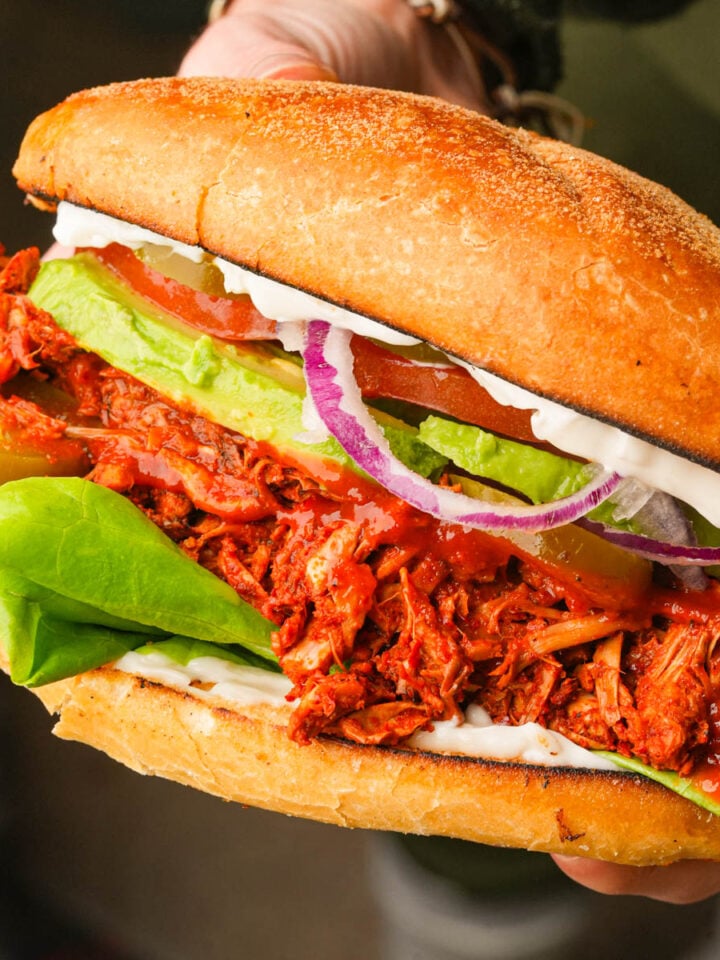 A close-up of a hand holding a large vegan torta adobada filled with shredded barbecue jackfruit, lettuce, avocado slices, tomato, red onion, pickles, and mayonnaise in a toasted bun.