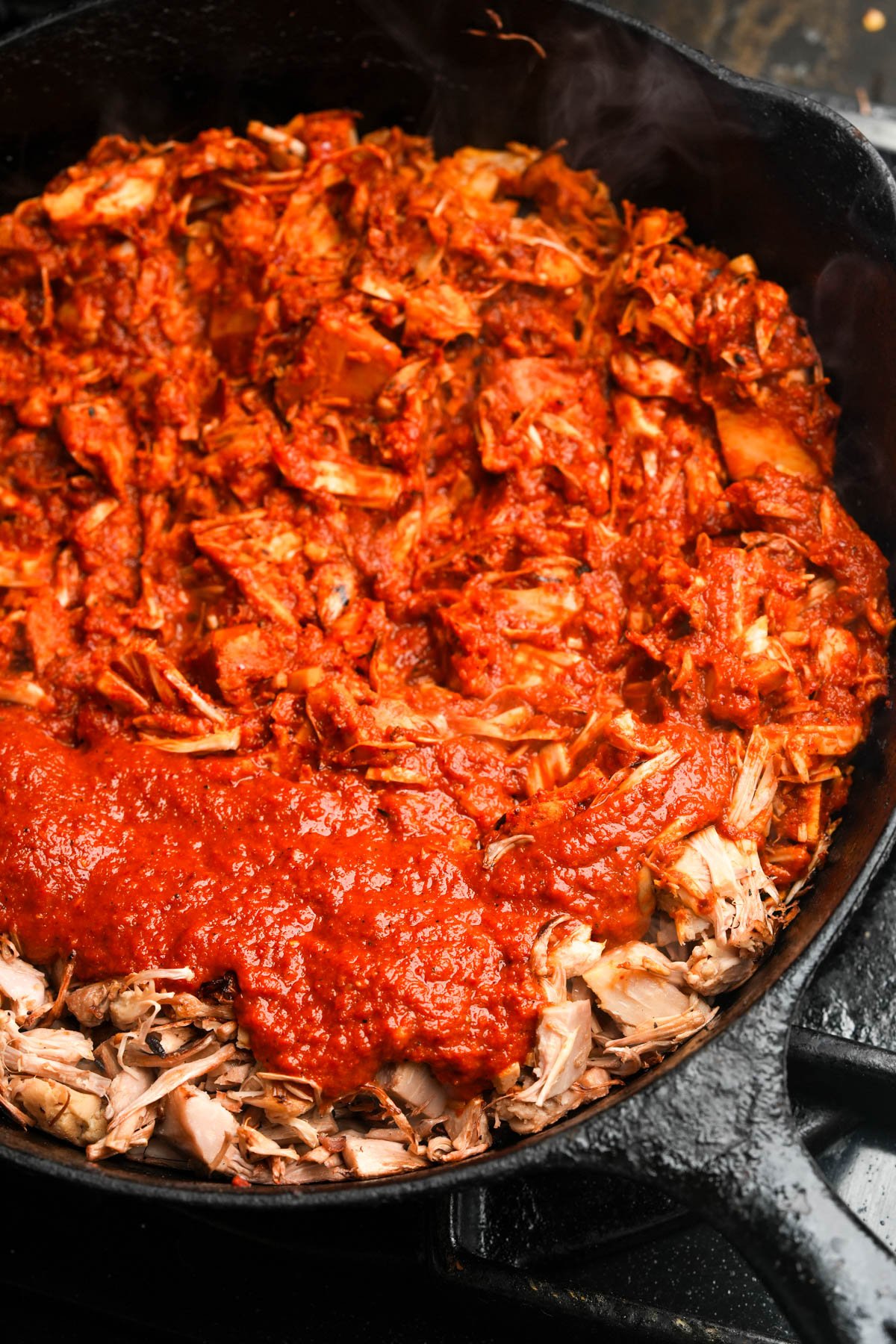 Shredded jackfruit in a cast iron skillet, partially covered with adobo sauce, with steam rising from the hot, cooked mixture.