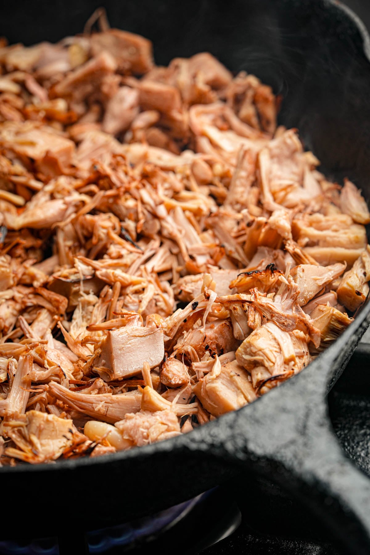 Close-up of shredded, cooked jackfruit in a black cast iron skillet, with visible steam rising, giving the appearance of pulled pork.