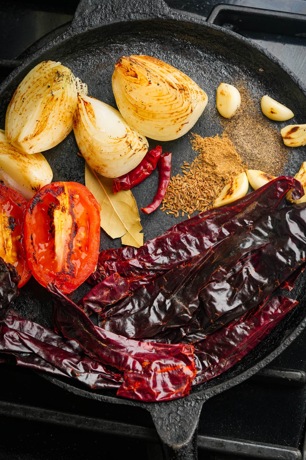 A cast iron skillet with charred onion, tomato, dried red chilies, bay leaf, garlic cloves, cumin seeds, dried chili peppers, and ground spices arranged and roasting together.