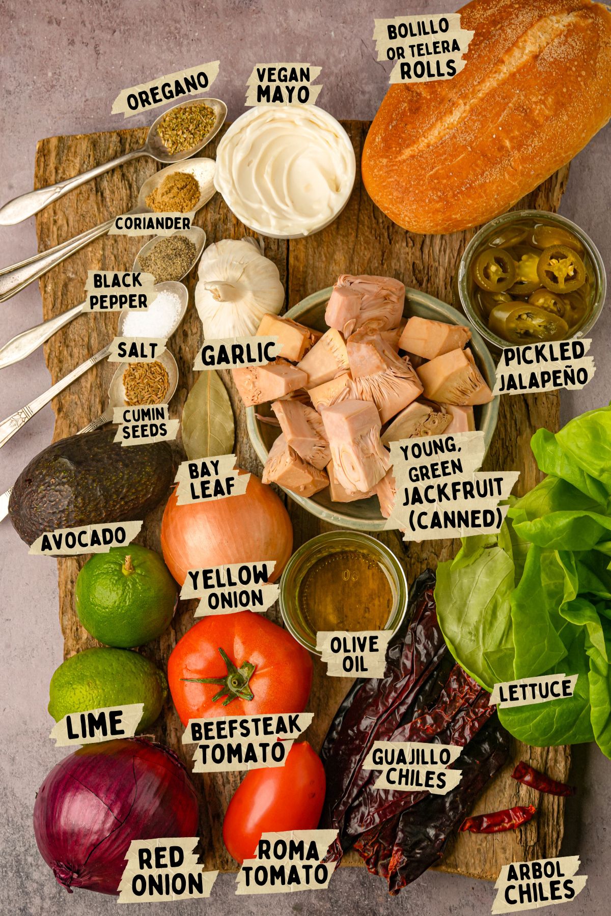 A wooden board displays labeled ingredients for vegan tortas adobada including jackfruit, bread roll, vegan mayo, spices, garlic, jalapeños, tomatoes, onions, avocado, lime, lettuce, pickled jalapeños, and dried chiles.