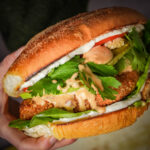 A hand holding a large vegan torta de milanesa filled with crispy fried tofu, leafy greens, pickles, tomato slices, white sauce, and a drizzle of orange sauce, all inside a toasted bun.