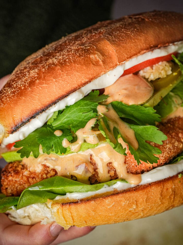 A hand holding a large vegan torta de milanesa filled with crispy fried tofu, leafy greens, pickles, tomato slices, white sauce, and a drizzle of orange sauce, all inside a toasted bun.