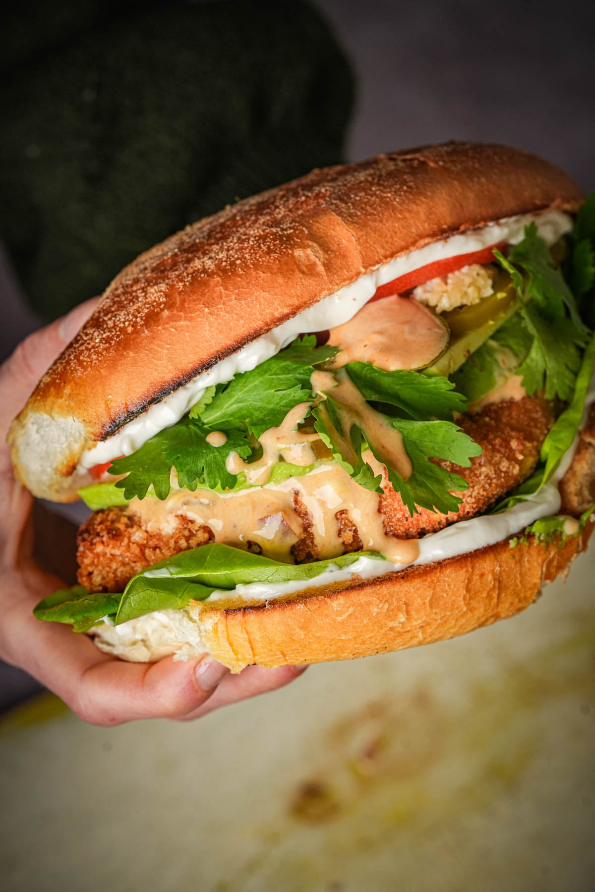 A hand holding a large vegan torta de milanesa filled with crispy fried tofu, leafy greens, sliced tomato, pickles, fresh cilantro, and creamy sauce in a toasted bun.