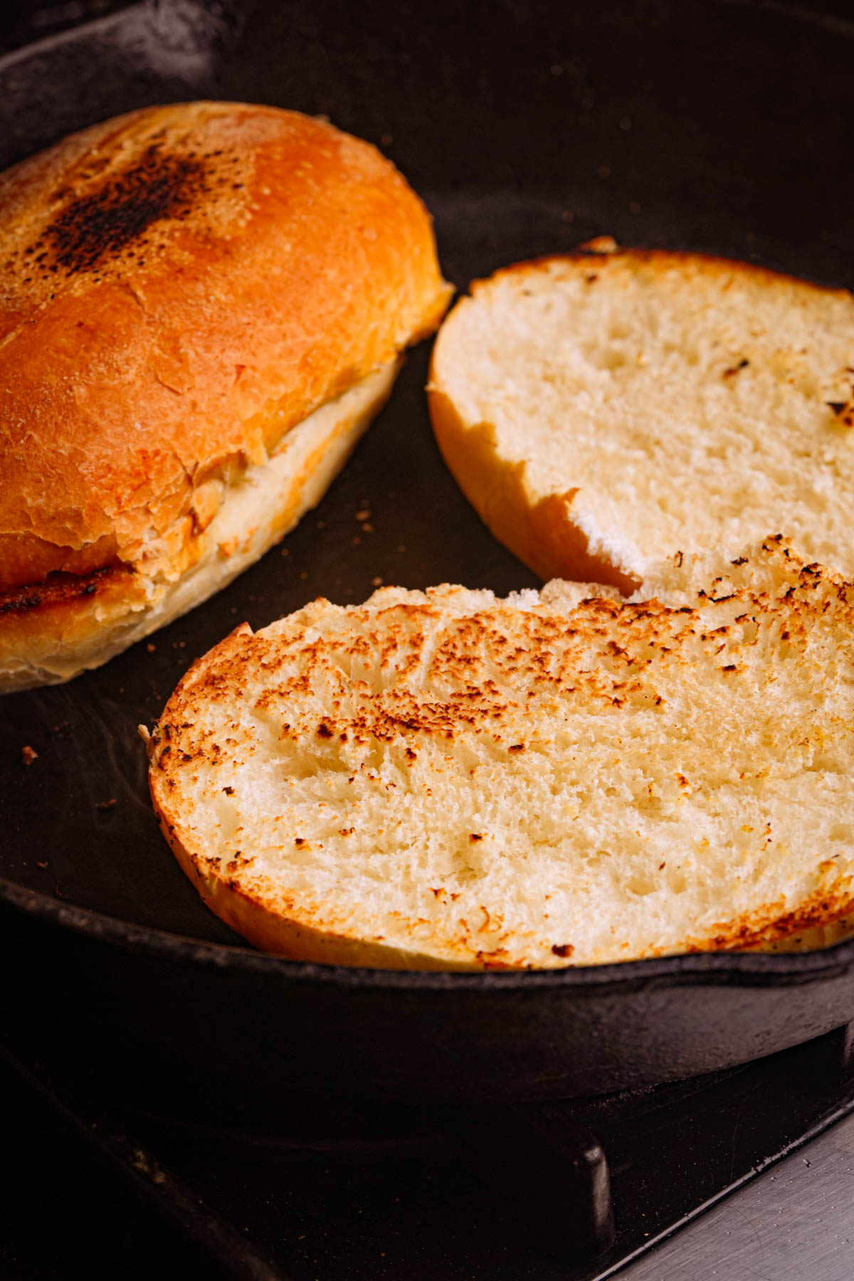 A sandwich bun sliced in half with the cut sides facing down, being toasted in a black skillet. One half is flipped to show a golden, crispy surface.