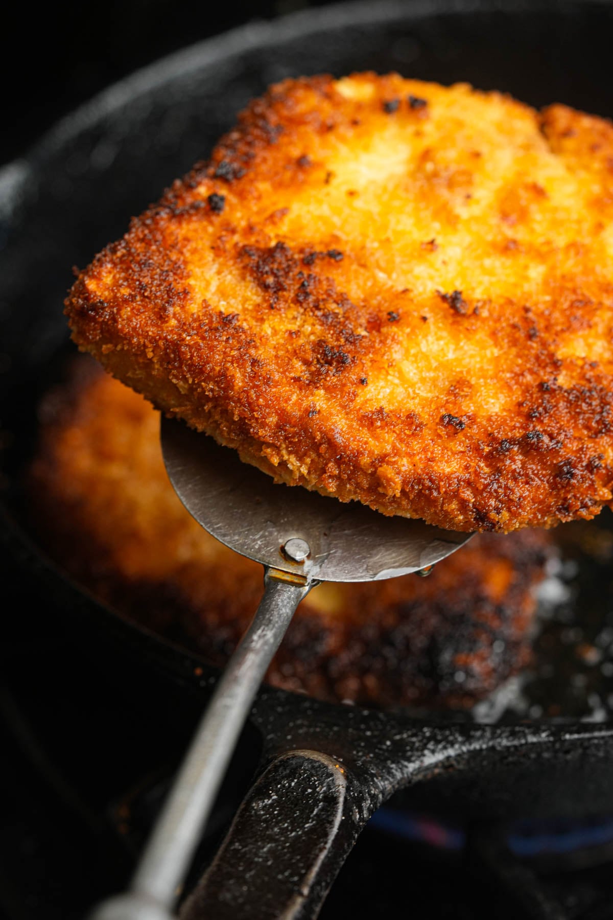A close-up of a golden-brown, crispy breaded tofu cutlet being lifted from a skillet with a metal spatula, showing its crunchy texture and cooked surface.