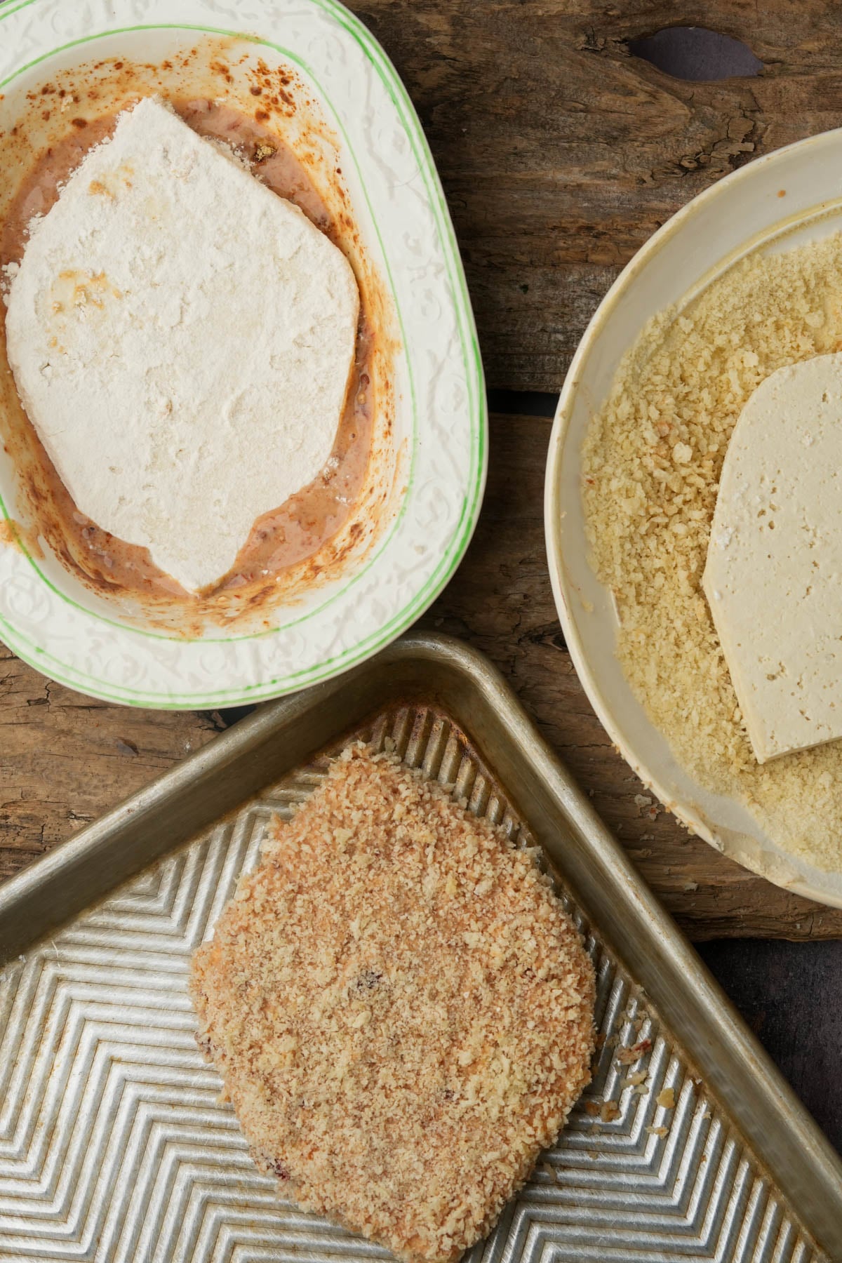 Three plates show stages of breading: tofu coated in flour, tofu dipped in batter, tofu covered in breadcrumbs on a metal baking tray, all on a rustic wooden surface.