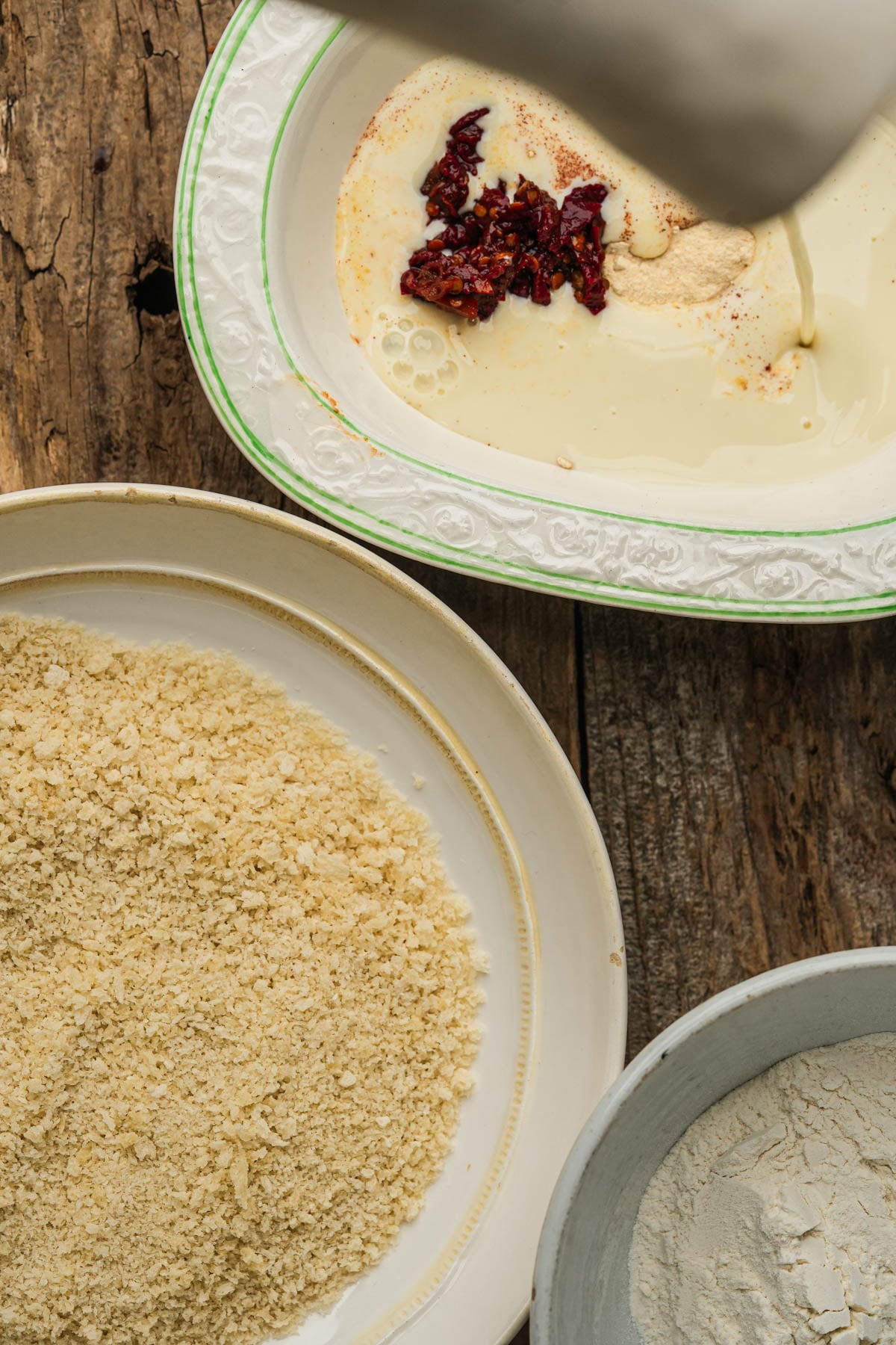Three bowls on a rustic wooden surface: one with flour, one with panko breadcrumbs, and one with a mixture of milk, spices, and chipotle chiles, with plant milk being poured in.