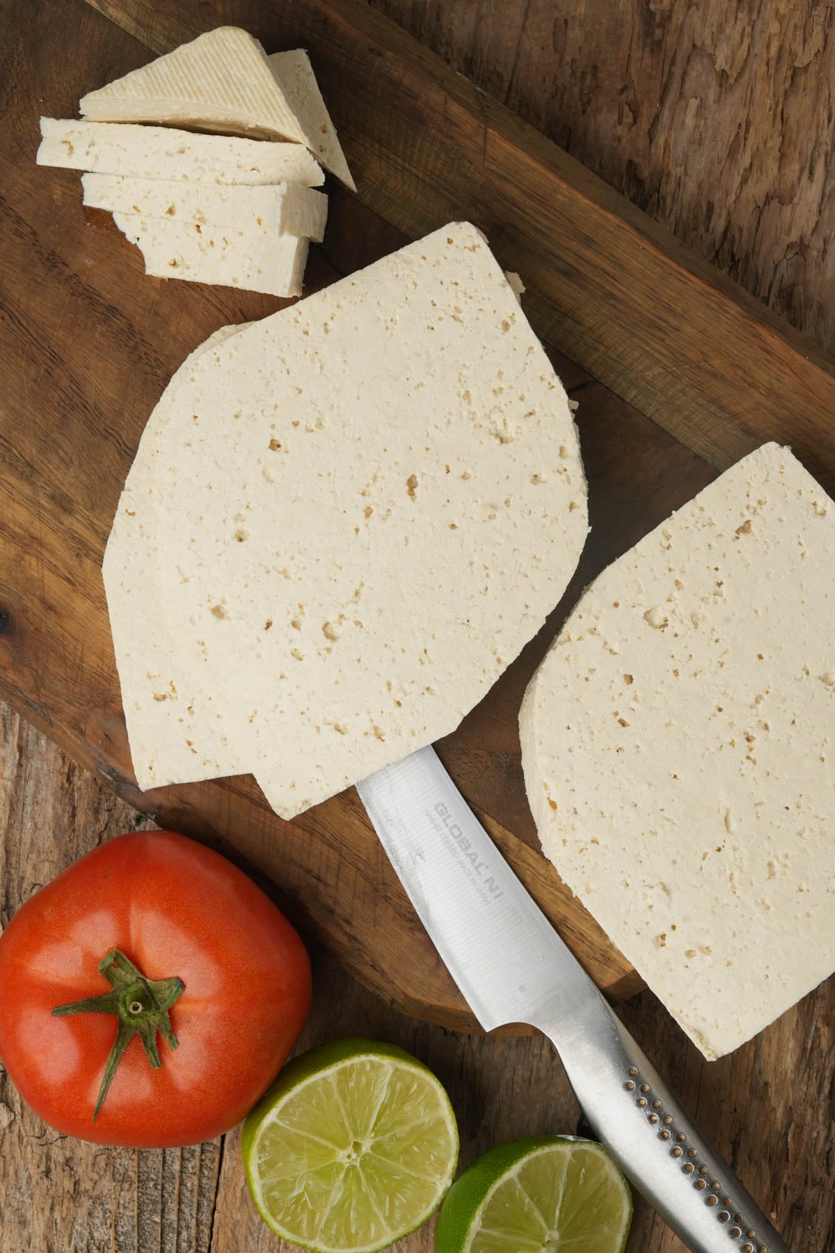 Two large slices and several small pieces of white tofu are on a wooden cutting board, with a knife, a whole tomato, and a halved lime and lemon next to them. The background is a rustic wooden surface.