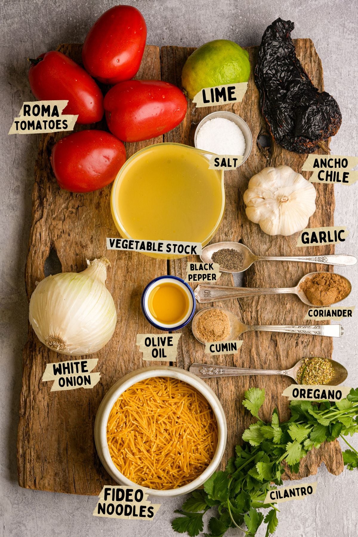 Ingredients for fideo soup arranged on a wooden board, including roma tomatoes, lime, ancho chile, garlic, salt, vegetable stock, white onion, black pepper, coriander, olive oil, cumin, oregano, fideo noodles, and cilantro.