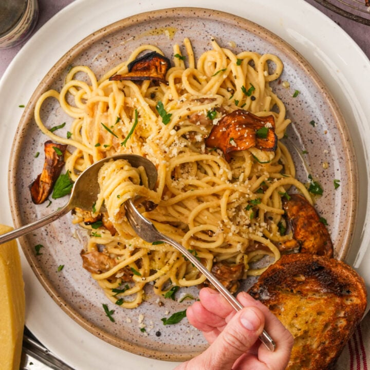 A hand uses a fork and spoon to twirl vegan carbonara with eggplant and parsley on a speckled plate, served with a slice of toasted bread.