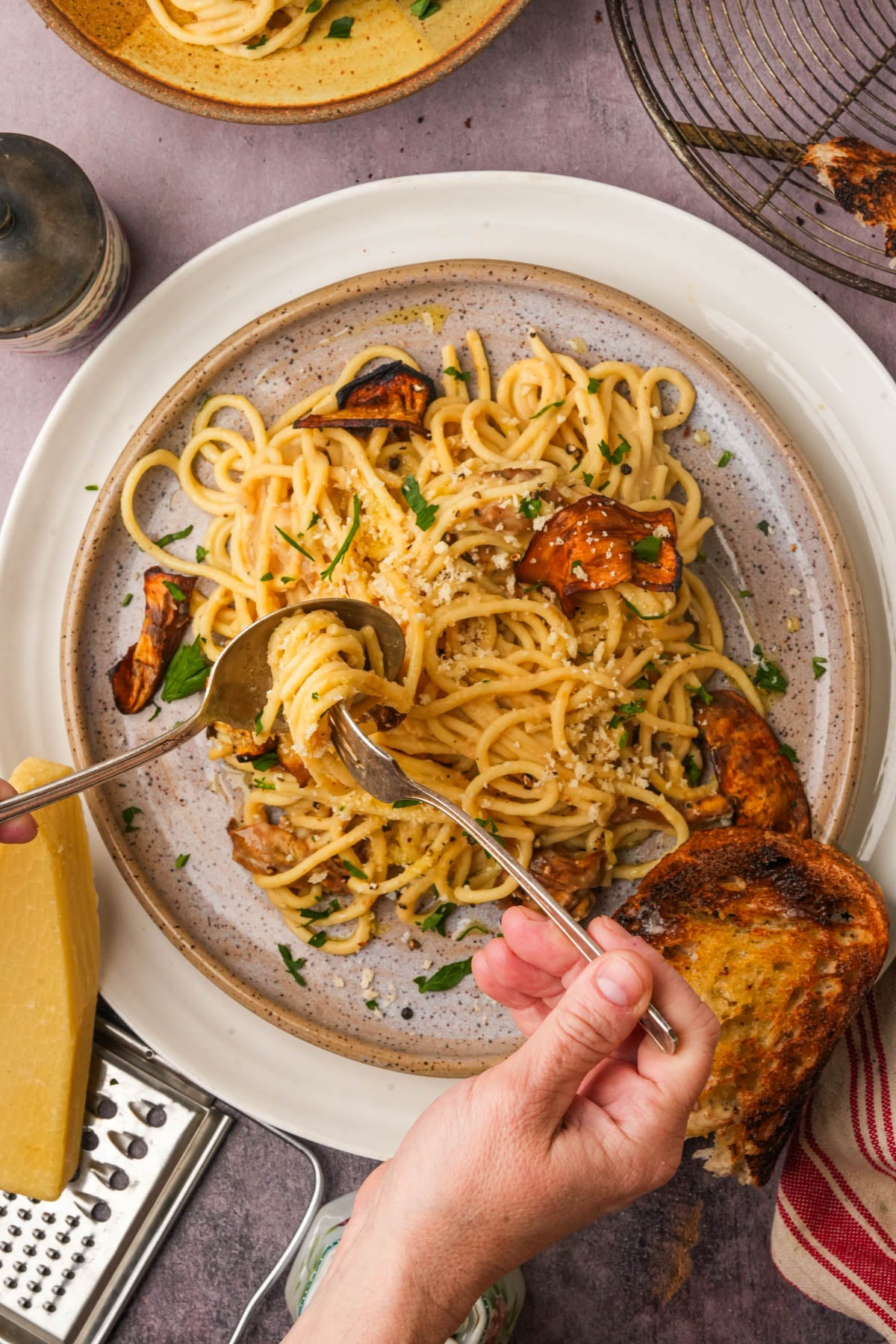 A plate of vegan carbonara with eggplant, garnished with parsley and grated cheese. A hand twirls pasta with a fork and spoon. Toasted bread and a cheese grater with a wedge of vegan cheese are nearby on the table.